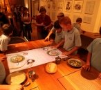 A group of school students look at a selection of props and AVs, including recreated bread and food in bowls. A large screen set into a table shows the arms of convicts eating food.