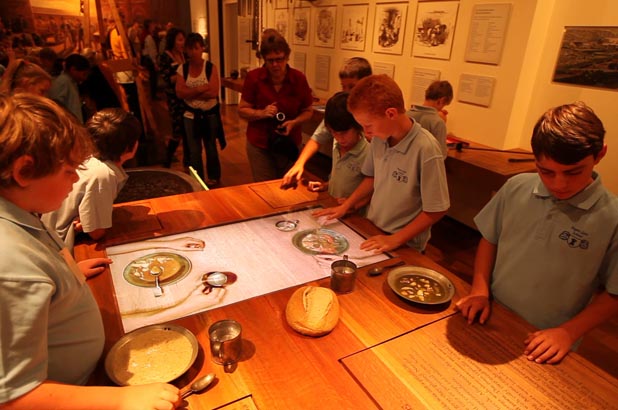 A group of school students look at a selection of props and AVs, including recreated bread and food in bowls. A large screen set into a table shows the arms of convicts eating food.