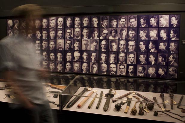 Image of a man moving past a wall covered in early 20th century mugshots. Underneath is a showcase containing weapons.